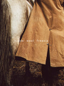 A close-up of a person wearing a tan suede and wool coat over a dark outfit, standing beside a horse outdoors. The text 'suede wool freesia' is overlaid across the center. A text overlay reading suede, wool and freesia as scent notes.