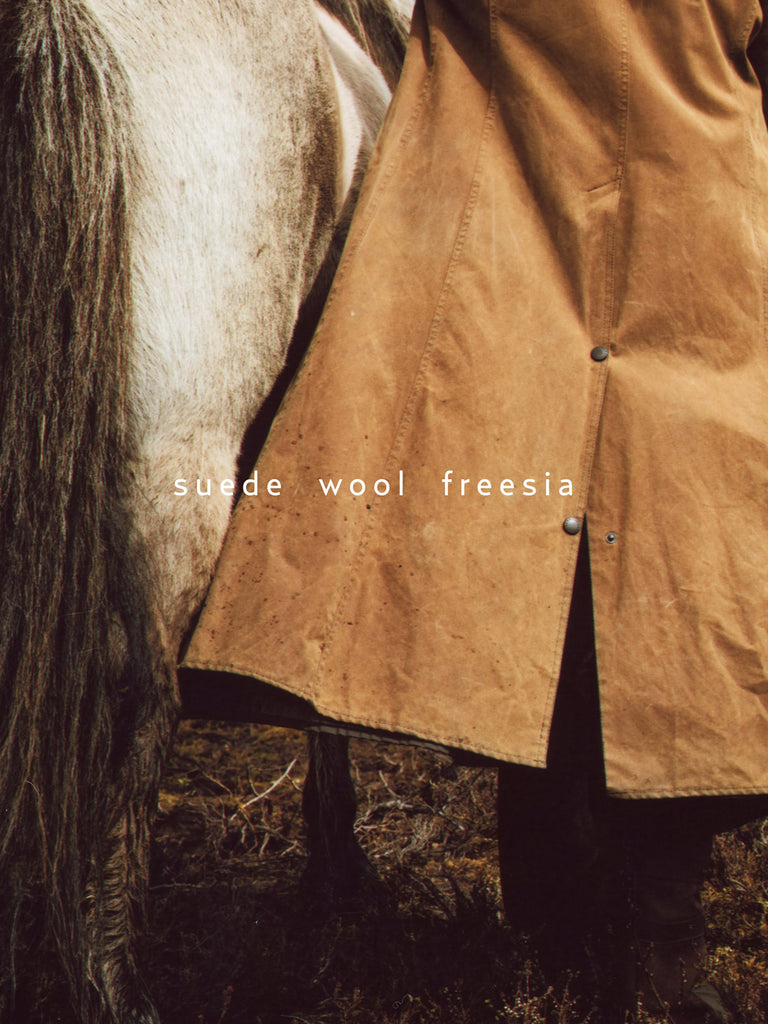 A close-up of a person wearing a tan suede and wool coat over a dark outfit, standing beside a horse outdoors. The text 'suede wool freesia' is overlaid across the center. A text overlay reading suede, wool and freesia as scent notes.