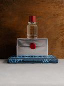 faluröd edp bottle with red cap on a white rectangular box with red seal, on top of a blue book with text, against a wooden background.