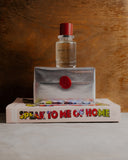 Never spring Perfume bottle, silver wrapped box with red seal, and book titled 'Speak to Me of Home' on a wooden surface.